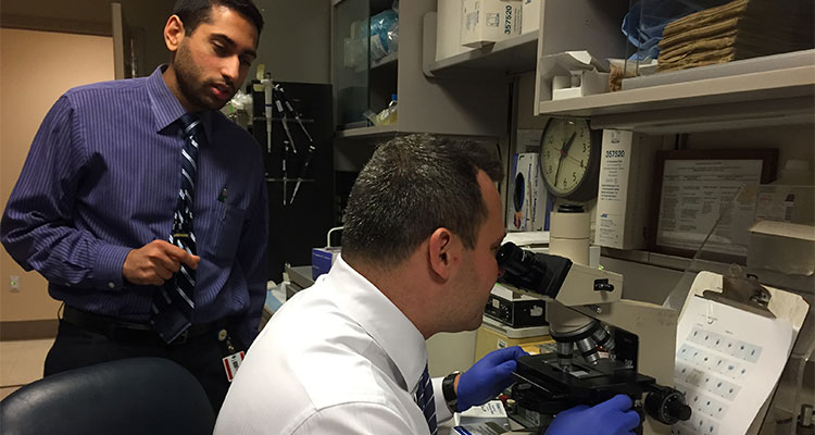 Male residents in lab. One works a microscope while the other stands and watches