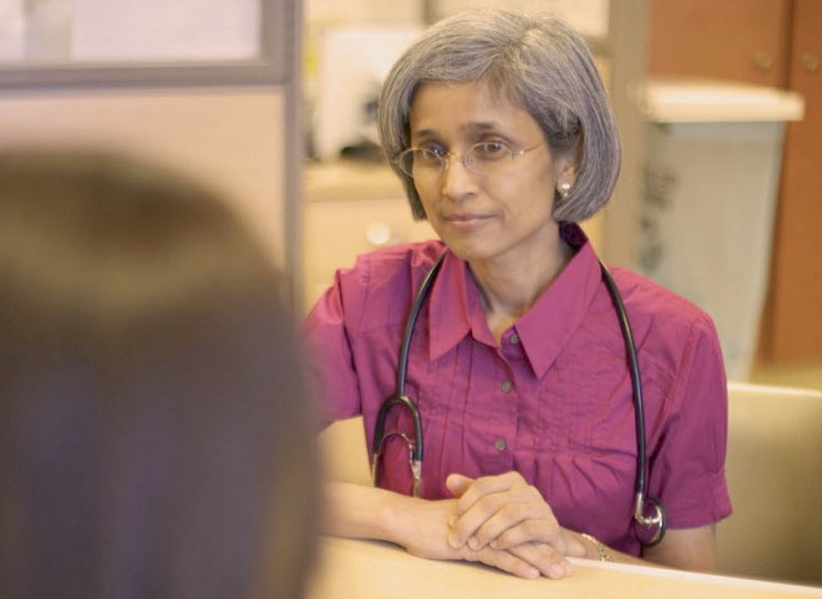 Female physician wearing purple button down shirt and stethoscope around her neck, talks to a patient